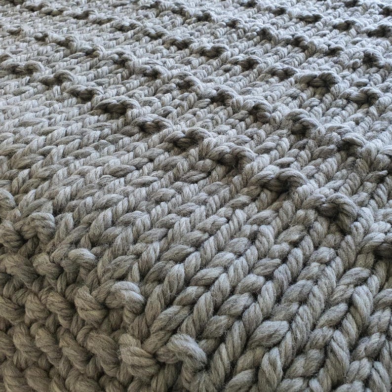 Close-up of a chunky knit blanket in a light gray color. The blanket features a textured pattern with raised, rounded stitches, creating a visually interesting surface. The thick yarn and tight weave suggest warmth and comfort.