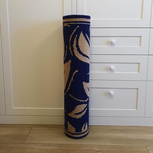 A rolled-up blue and beige patterned rug with a floral design. The rug is rolled up and standing upright on a wooden floor.
