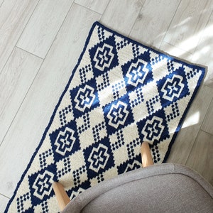 May include: A white and blue crocheted rug with a geometric pattern. The rug has a diamond shape with a cross in the center of each diamond. The rug is on a light wood floor.