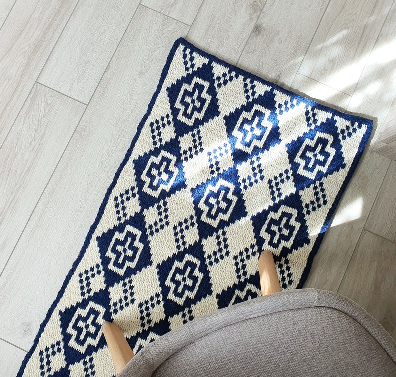 May include: A white and blue crocheted rug with a geometric pattern. The rug has a diamond shape with a cross in the center of each diamond. The rug is on a light wood floor.