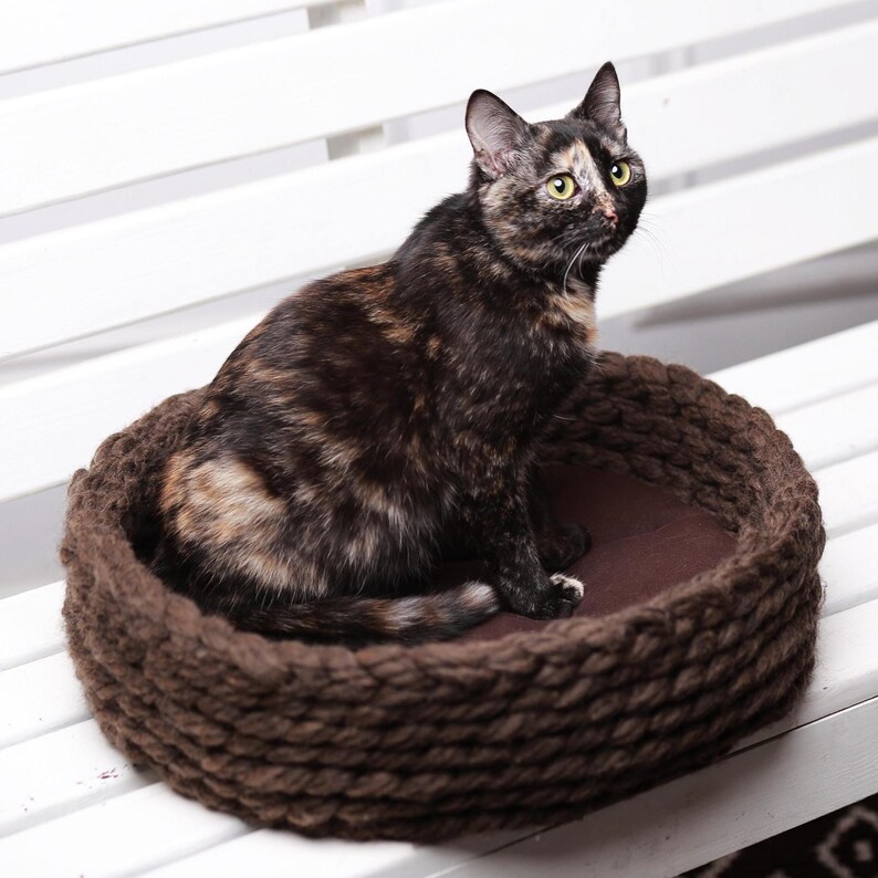 May include: A tortoiseshell cat with black, brown, and orange fur sits inside a round, dark brown, thick-knit yarn pet bed. The bed features a textured, braided rope-like construction with a solid dark brown fabric base cushion inside.