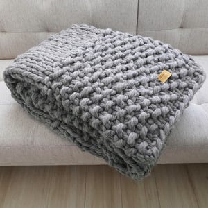 A folded, chunky knit blanket in a light gray color. The blanket is made of thick yarn and has a textured appearance. A small, rectangular tag is attached to the blanket.