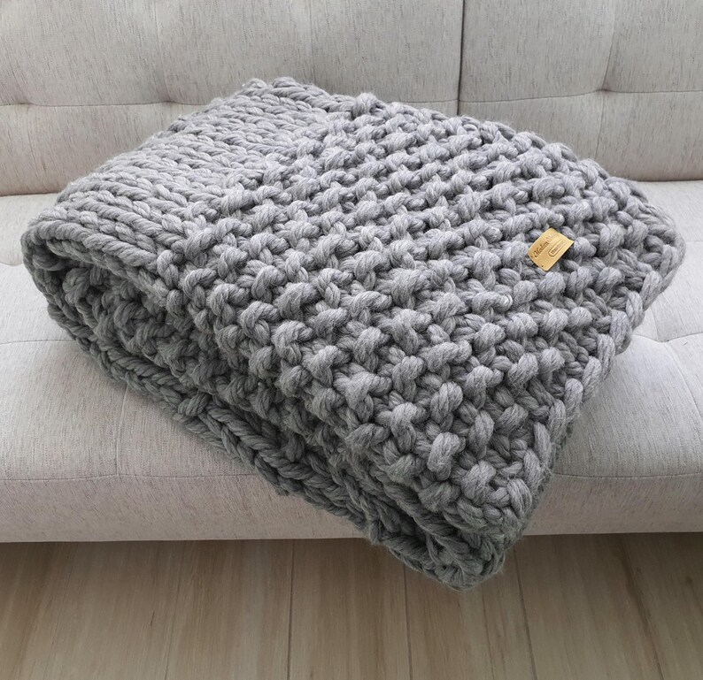 A folded, chunky knit blanket in a light gray color. The blanket is made of thick yarn and has a textured appearance. A small, rectangular tag is attached to the blanket.