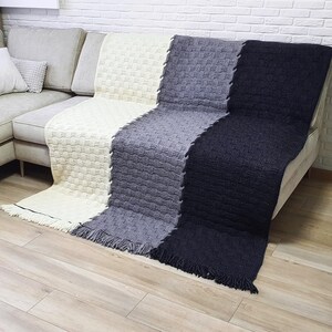 A woven throw blanket with three sections in cream, gray, and black. The blanket has a textured weave and fringe on the edges. The blanket is draped over a couch.