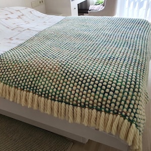 A textured, hand-knitted blanket in shades of green, white, and tan, draped over a bed. The blanket features a bobble stitch pattern and a cream-colored fringe along the bottom edge. The blanket is a cozy home decor item.