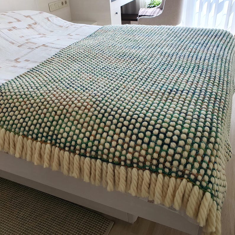 A textured, hand-knitted blanket in shades of green, white, and tan, draped over a bed. The blanket features a bobble stitch pattern and a cream-colored fringe along the bottom edge. The blanket is a cozy home decor item.
