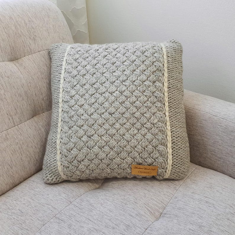 May include: A square, light grey knitted cushion with a woven pattern. It features a cream-coloured border and a small, rectangular brown tag. The cushion is placed on a light grey sofa.