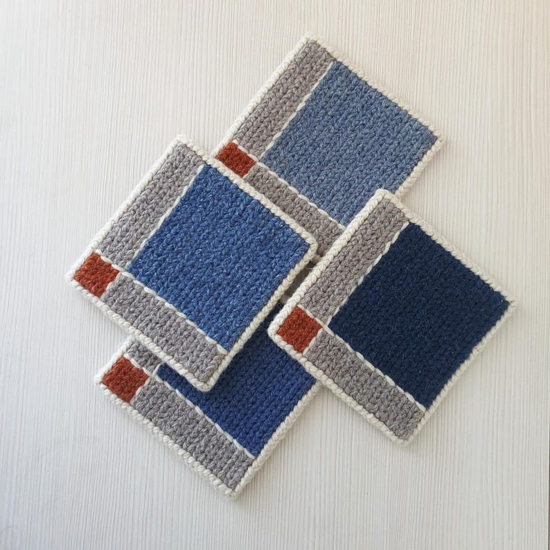 K&ouml;nnte beinhalten: Vier quadratische H&auml;keluntersetzer in Blau-, Grau- und Rostt&ouml;nen. Jeder Untersetzer hat ein zentrales blaues Quadrat, einen grauen Rand und einen rostfarbenen Akzent. Die Untersetzer sind auf einer wei&szlig;en Oberfl&auml;che angeordnet.