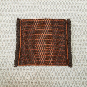 Puede incluir: Una alfombra cuadrada tejida marr&oacute;n y naranja con un patr&oacute;n de panal. La alfombra tiene un borde marr&oacute;n oscuro con flecos. El fondo es una tela color crema con un patr&oacute;n repetitivo.