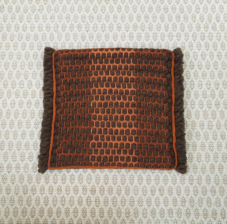 Puede incluir: Una alfombra cuadrada tejida marr&oacute;n y naranja con un patr&oacute;n de panal. La alfombra tiene un borde marr&oacute;n oscuro con flecos. El fondo es una tela color crema con un patr&oacute;n repetitivo.