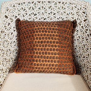 Puede incluir: Un coj&iacute;n decorativo cuadrado con un patr&oacute;n tejido marr&oacute;n y naranja. El coj&iacute;n tiene un borde con flecos y est&aacute; colocado sobre una superficie de color claro. El coj&iacute;n est&aacute; sobre una silla de mimbre blanca.