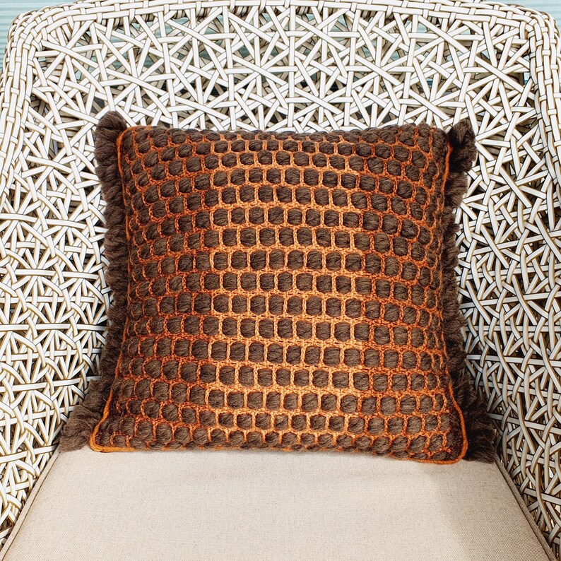 Puede incluir: Un coj&iacute;n decorativo cuadrado con un patr&oacute;n tejido marr&oacute;n y naranja. El coj&iacute;n tiene un borde con flecos y est&aacute; colocado sobre una superficie de color claro. El coj&iacute;n est&aacute; sobre una silla de mimbre blanca.