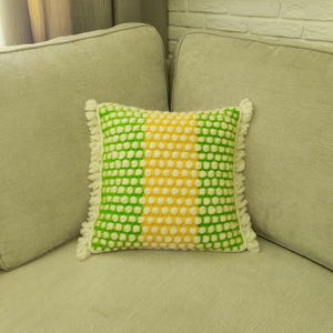 May include: A square decorative pillow with a textured pattern in green, yellow, and white. The pillow has a fringe trim and is placed on a light-colored sofa. The pillow's design features a central yellow stripe flanked by green sections.