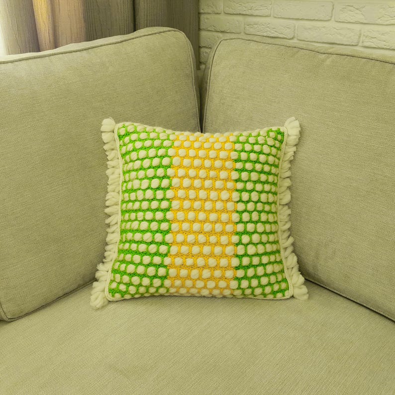 May include: A square decorative pillow with a textured pattern in green, yellow, and white. The pillow has a fringe trim and is placed on a light-colored sofa. The pillow's design features a central yellow stripe flanked by green sections.