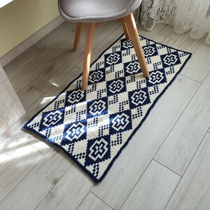 May include: A white and navy blue geometric patterned rug with a diamond design. The rug is made of a soft, woven material and has a subtle sheen.