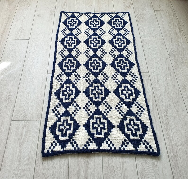 May include: A white and navy blue crocheted rug with a geometric pattern. The rug has a diamond shape with a cross in the center of each diamond.