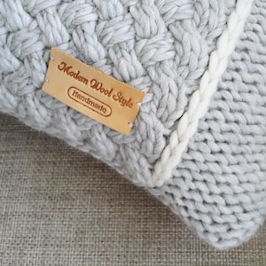 May include: A close-up shot of a light grey, handmade knitted cushion with a woven pattern. A white knitted border runs along the edge. A brown leather tag reads "Modern Wool Style Handmade".