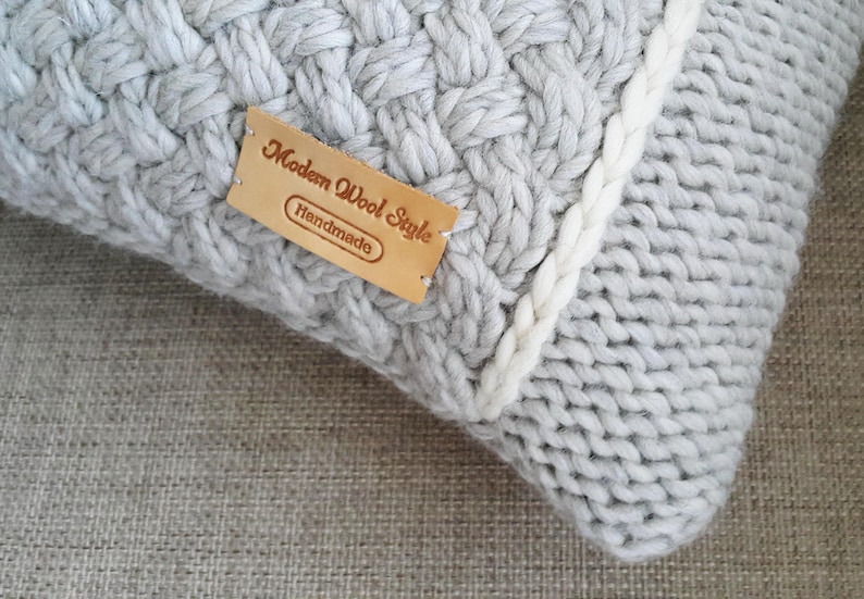 May include: A close-up shot of a light grey, handmade knitted cushion with a woven pattern. A white knitted border runs along the edge. A brown leather tag reads "Modern Wool Style Handmade".