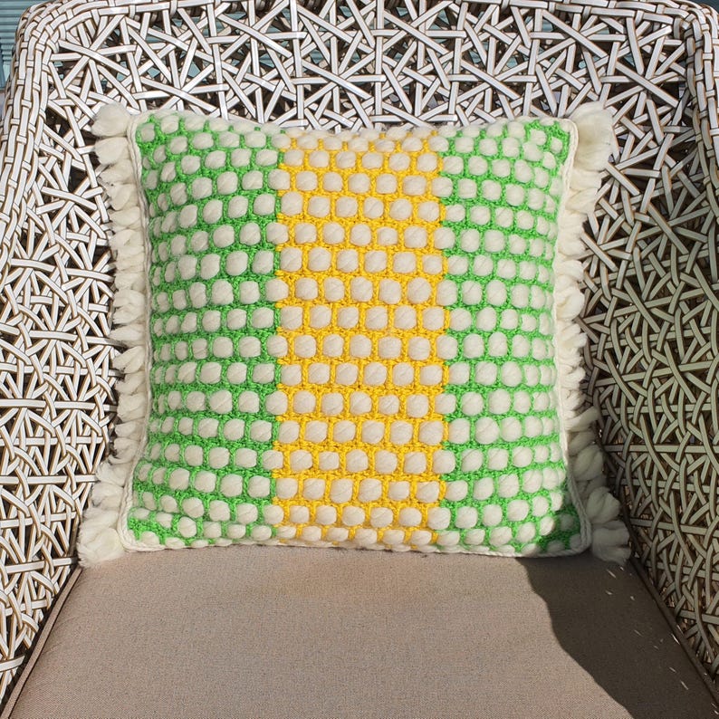 May include: A square decorative pillow with a textured pattern of white, green, and yellow. The pillow has a fringe trim and is placed on a neutral-colored surface. The pillow is on a white wicker chair.