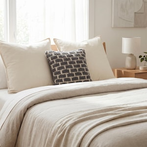 May include: A bed dressed in cream-coloured bedding, including pillows and a duvet. A decorative grey and white textured cushion sits centrally. A small lamp and artwork are visible in the background, suggesting a bedroom setting.