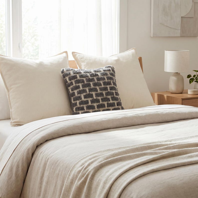 May include: A bed dressed in cream-coloured bedding, including pillows and a duvet. A decorative grey and white textured cushion sits centrally. A small lamp and artwork are visible in the background, suggesting a bedroom setting.