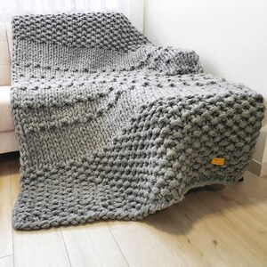 A chunky knit blanket in varying shades of gray, draped over a sofa. The blanket features a large, textured stitch pattern and a soft, inviting appearance. The blanket is made of a thick yarn, creating a cozy and warm aesthetic.