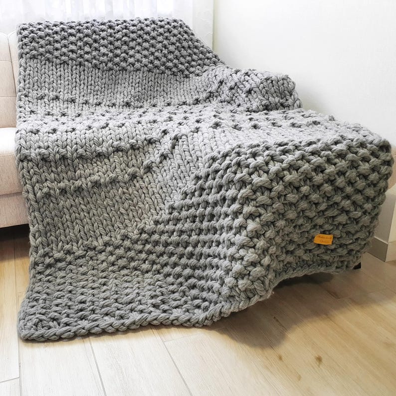 A chunky knit blanket in varying shades of gray, draped over a sofa. The blanket features a large, textured stitch pattern and a soft, inviting appearance. The blanket is made of a thick yarn, creating a cozy and warm aesthetic.