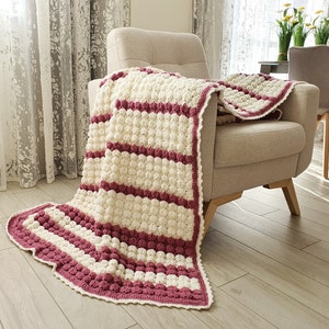A crocheted throw blanket with white and burgundy stripes. The blanket is draped over a light brown armchair.