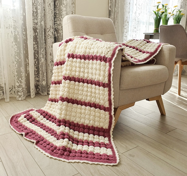 A crocheted throw blanket with white and burgundy stripes. The blanket is draped over a light brown armchair.