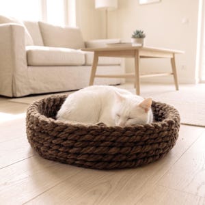 A white cat sleeping curled up inside a round brown chunky knit wool pet bed, placed on a light hardwood floor in a bright Scandinavian-style living room with a cream sofa and wooden coffee table in the background.