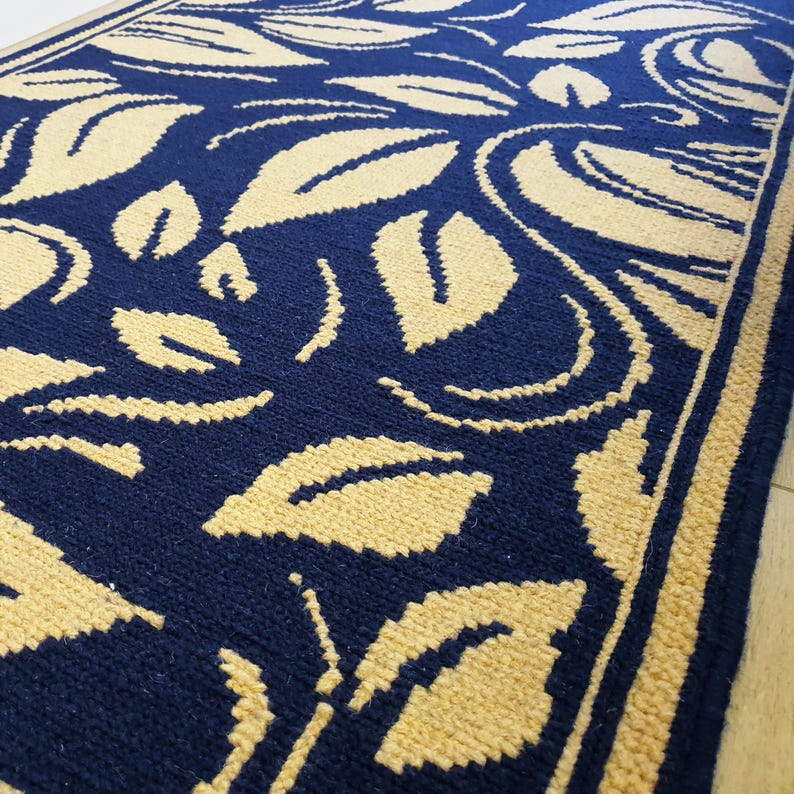 Pu&ograve; includere: Un tappeto blu navy e beige a motivi floreali. Il tappeto presenta un motivo ripetuto di foglie e viti stilizzate in beige su uno sfondo blu scuro. I bordi sono bordati con una striscia beige.