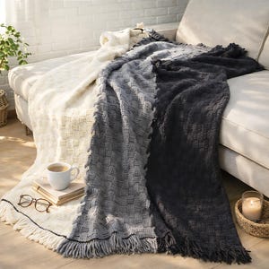 A three-toned knitted throw blanket with fringe. The blanket is white, gray, and black. It is draped over a couch.