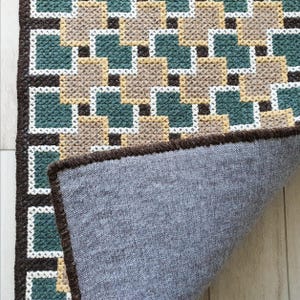 A brown and green geometric patterned rug with a gray backing. The rug is folded in half, showing the backing. Back side of handmade wool rug showing structure and craftsmanship, Beige, Brown, Green and Teal threads visible