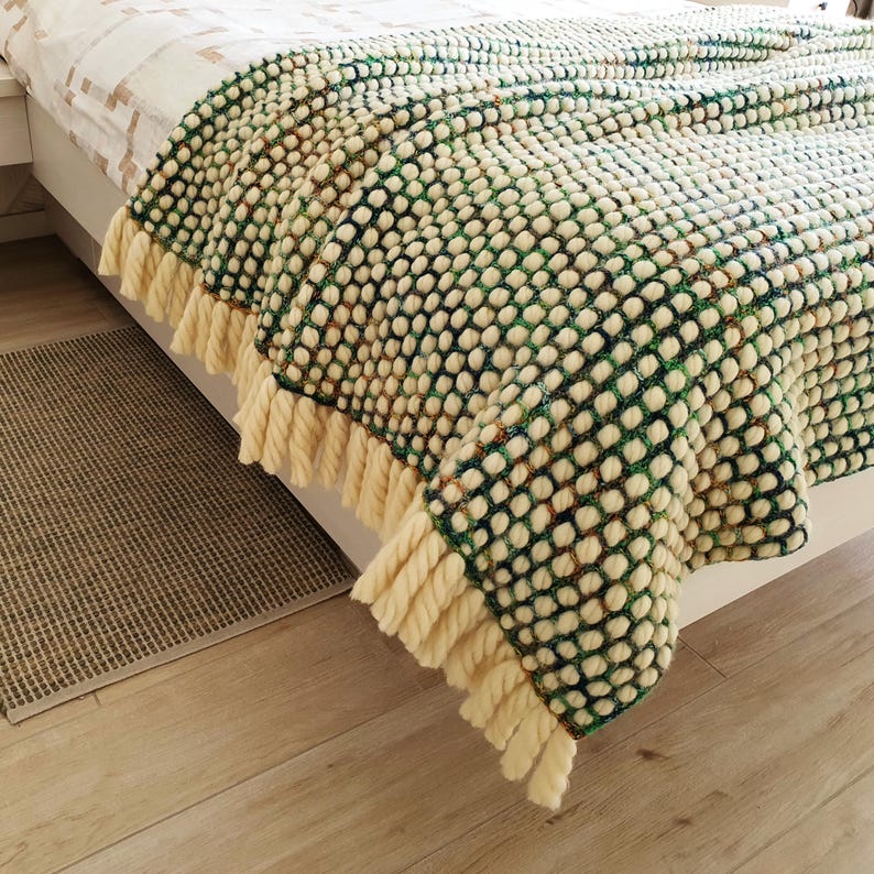 A textured, cream-colored blanket with a repeating dot pattern in green and navy blue. The blanket has a thick, cream-colored fringe along the bottom edge. The blanket is draped over a bed, showcasing its cozy design.