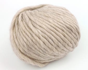 May include: A ball of thick, beige yarn. The yarn is tightly wound, with visible strands creating a textured surface. The neutral color suggests it could be used for various knitting or crochet projects, such as blankets or scarves.