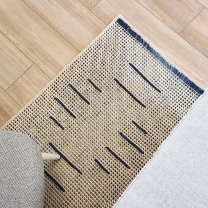 May include: A beige and black woven rug with a geometric pattern. The rug has a fringe border and is made of natural fibers.