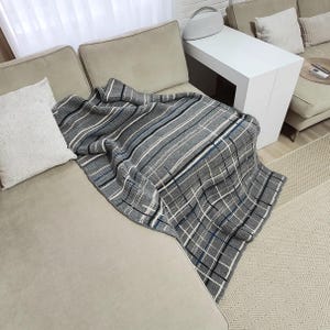 A gray and white plaid throw blanket draped over a light gray couch with two white throw pillows. The couch is in a living room setting with a white side table and a rug.