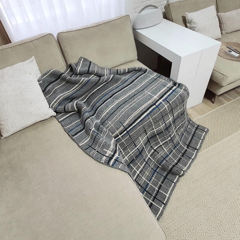 A gray and white plaid throw blanket draped over a light gray couch with two white throw pillows. The couch is in a living room setting with a white side table and a rug.