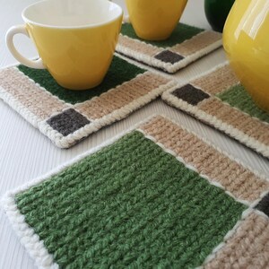 May include: A set of four handmade crochet coasters. Each coaster features a green, tan, and gray square design with a white border. Two yellow mugs are visible, resting on two of the coasters. The coasters are designed to protect surfaces from heat and moisture.
