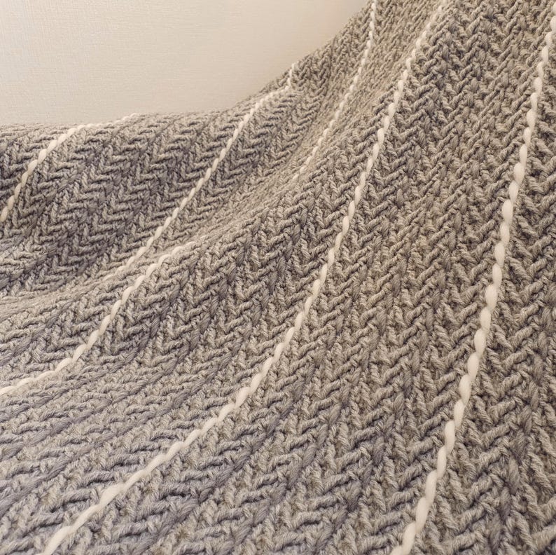A gray and white woven throw blanket with a herringbone pattern. The blanket has a soft, textured feel.