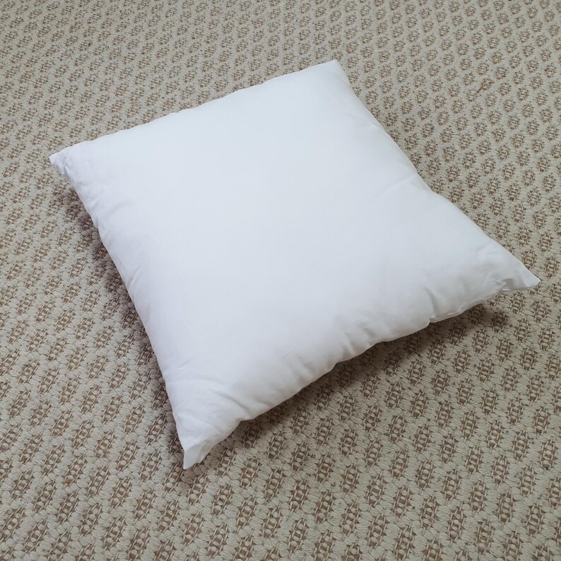 May include: A square, white pillow on a patterned beige carpet. The pillow is fluffy and appears to be made of a soft material. The pillow is centered in the image and takes up most of the frame.