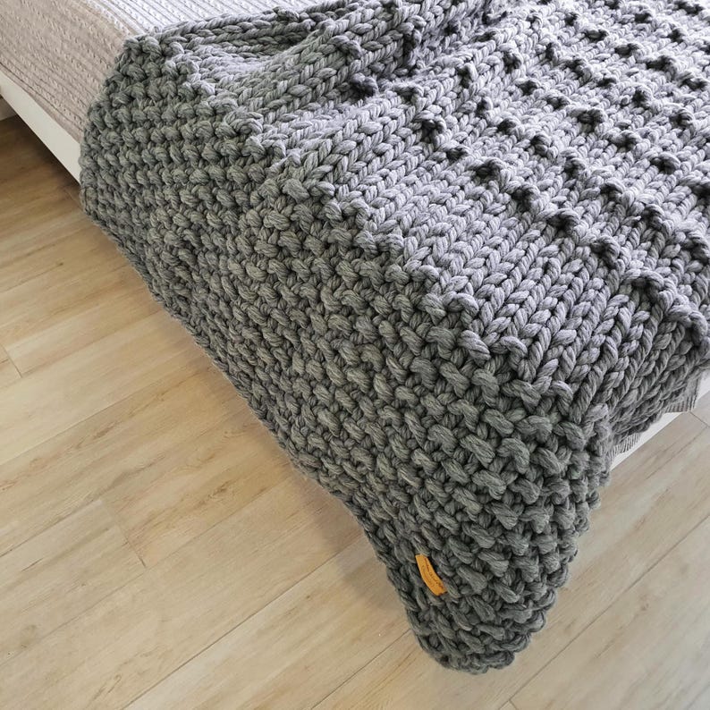 A chunky knit blanket in a soft gray color. The blanket has a textured, raised pattern and is draped over a bed. A small, yellow tag is attached to the corner of the blanket. The blanket is made of thick yarn.