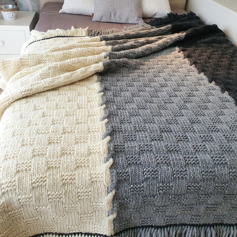 A knitted throw blanket with a cream and gray checkered pattern. The blanket has a fringe border and is folded on a bed.