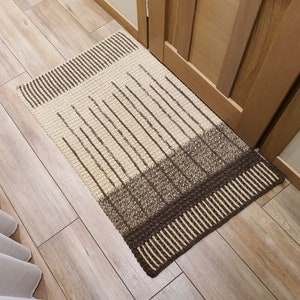 May include: A brown and beige striped crocheted rug with a geometric pattern. The rug is placed in front of a wooden door.