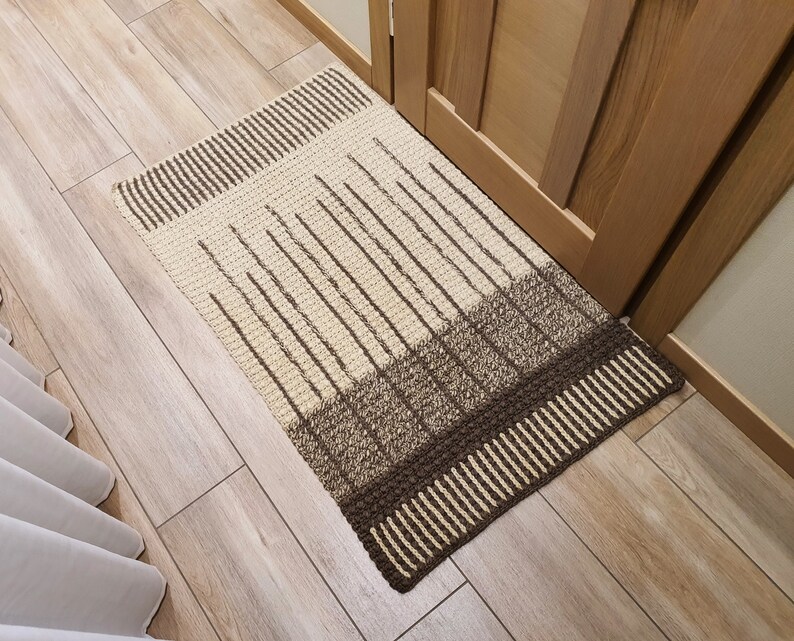 May include: A brown and beige striped crocheted rug with a geometric pattern. The rug is placed in front of a wooden door.
