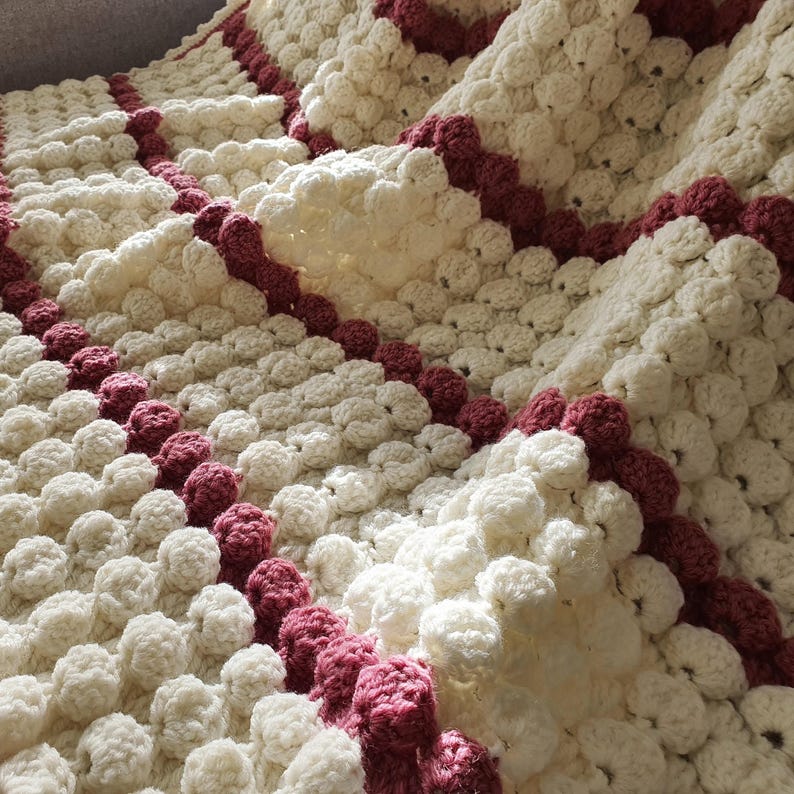 A white crocheted blanket with a textured, puffy design. The blanket has burgundy stripes running across the width of the blanket.