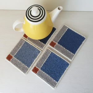 K&ouml;nnte beinhalten: Eine gelbe Teekanne mit einem schwarz-wei&szlig;en kreisf&ouml;rmigen Design auf dem Deckel steht auf vier quadratischen, geh&auml;kelten Untersetzern. Die Untersetzer haben eine blaue Mitte, einen grauen Rand und einen kleinen braunen quadratischen Akzent. Die Untersetzer sind etwa 10 cm gro&szlig;.