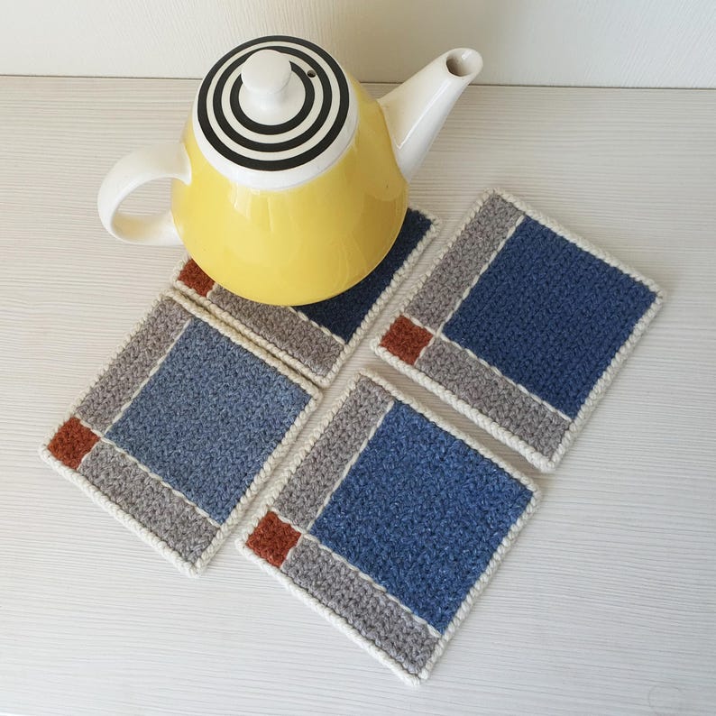K&ouml;nnte beinhalten: Eine gelbe Teekanne mit einem schwarz-wei&szlig;en kreisf&ouml;rmigen Design auf dem Deckel steht auf vier quadratischen, geh&auml;kelten Untersetzern. Die Untersetzer haben eine blaue Mitte, einen grauen Rand und einen kleinen braunen quadratischen Akzent. Die Untersetzer sind etwa 10 cm gro&szlig;.