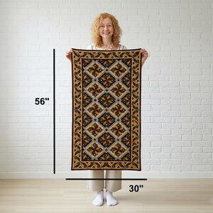 A rectangular rug with a geometric pattern in shades of brown, gold, and black. The rug features a repeating diamond and floral design, with a dark brown border. The rug is held up against a white brick wall.