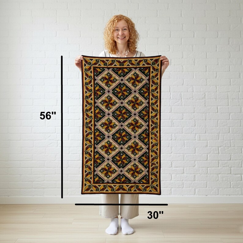 A rectangular rug with a geometric pattern in shades of brown, gold, and black. The rug features a repeating diamond and floral design, with a dark brown border. The rug is held up against a white brick wall.